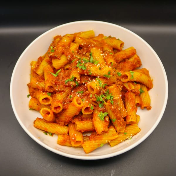 A white bowl filled with rigatoni pasta in a rich red sauce, garnished with parsley and cheese.