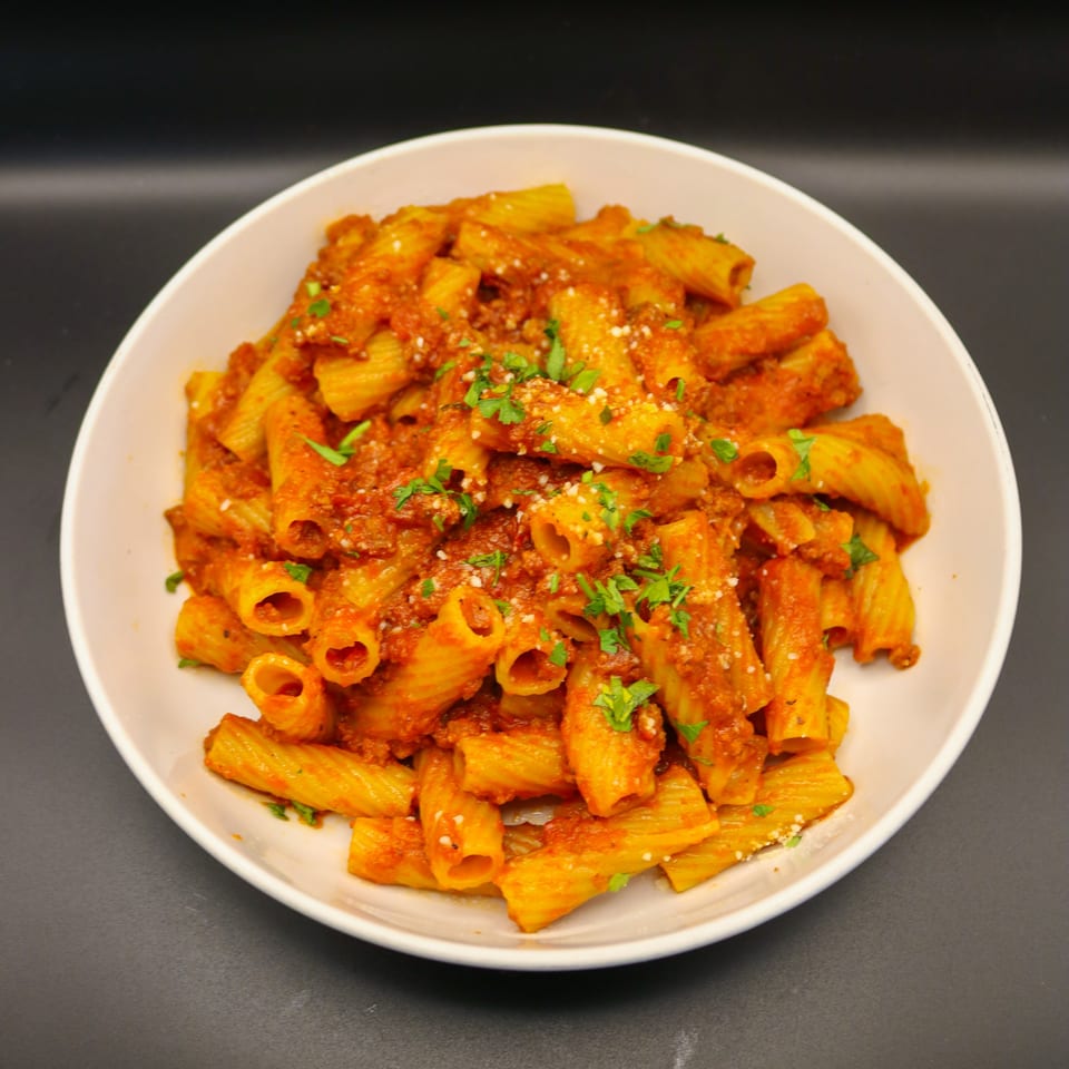 A white bowl filled with rigatoni pasta in a rich red sauce, garnished with parsley and cheese.