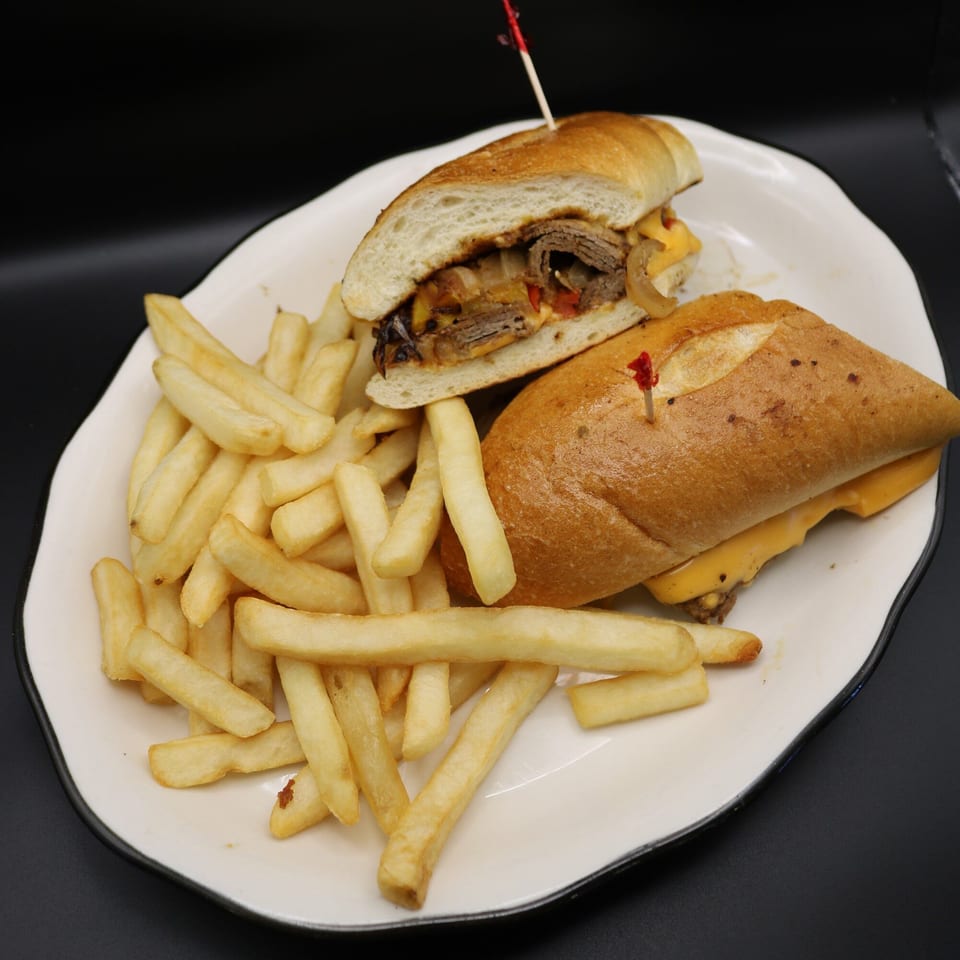 A Johnson Diner & Bar cheesesteak sandwich with fries on a white plate against a dark background.