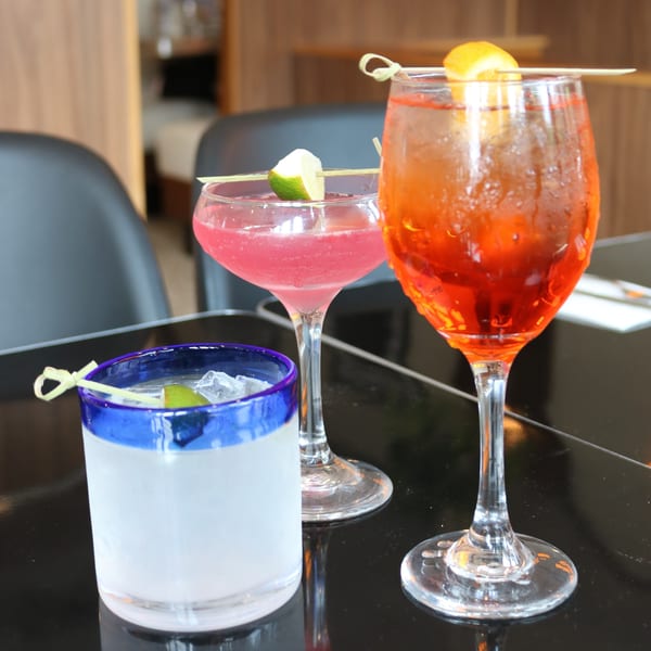 Three colorful cocktails, including a pink martini and an orange spritz, on a dark table.