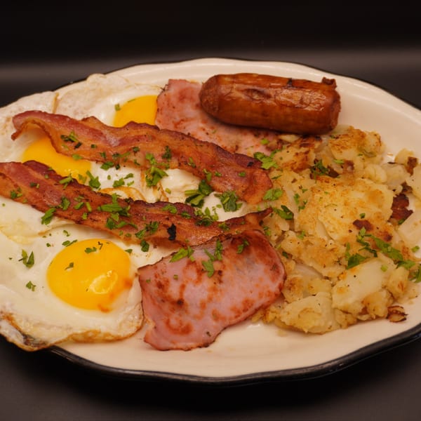 A breakfast platter with fried eggs, crispy bacon, ham, sausage, and home fries, garnished with parsley.