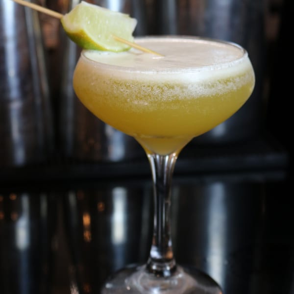 A Johnson Diner & Bar cocktail, yellow with foam, garnished with a lime wedge on a dark bar.
