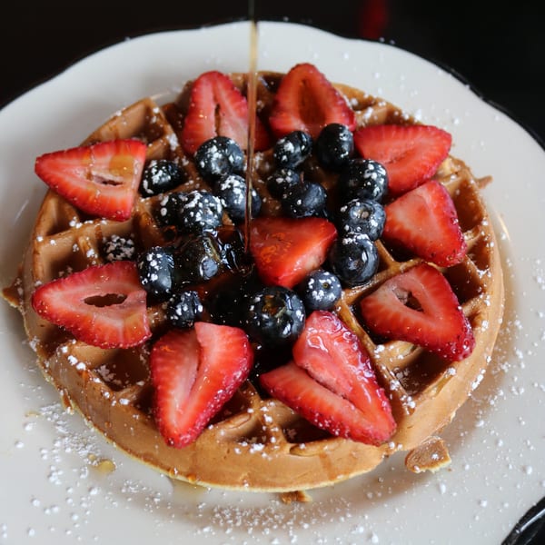 Golden waffle topped with fresh strawberries, blueberries, powdered sugar, and maple syrup pouring.